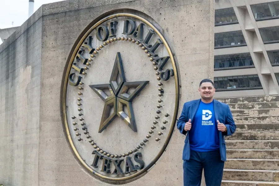 Dallas College alum Jaime Resendez stands outdoors in front of a City of Dallas building, wearing business-casual attire and posing confidently on a walkway.
