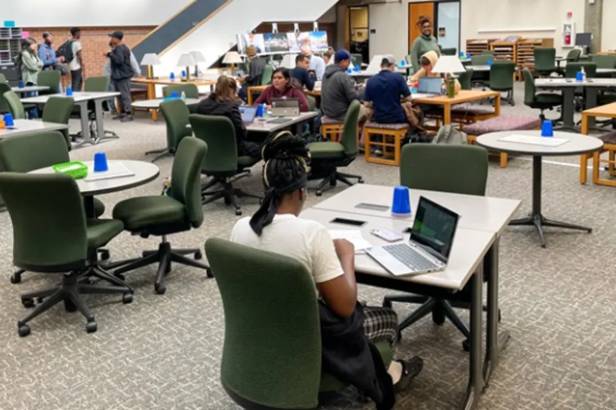 A library with about 16 people, relatively low green chairs, and a variety of circular and rectangular tables.