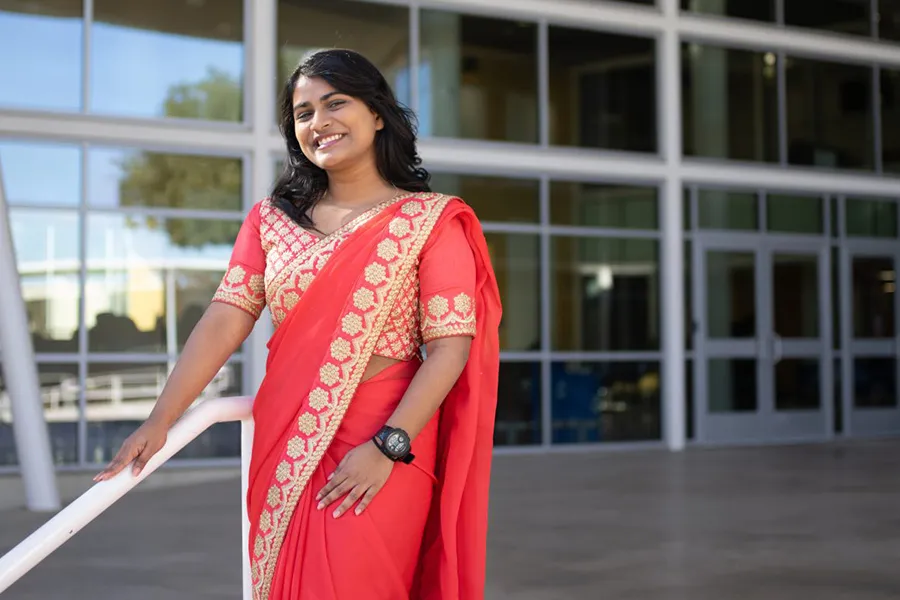 Tiffiya standing beside a white handrail in front of a building on a Dallas College campus. She is wearing a red sari with a gold pattern. A dark wristwatch is visible on one wrist.