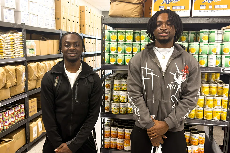 Two students stand in front of campus food pantry shelves.