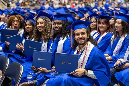 A group of Dallas College students hold up their diplomas at their graduation ceremony.