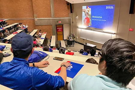 Students sit in a large auditorium facing a screen showing a presentation