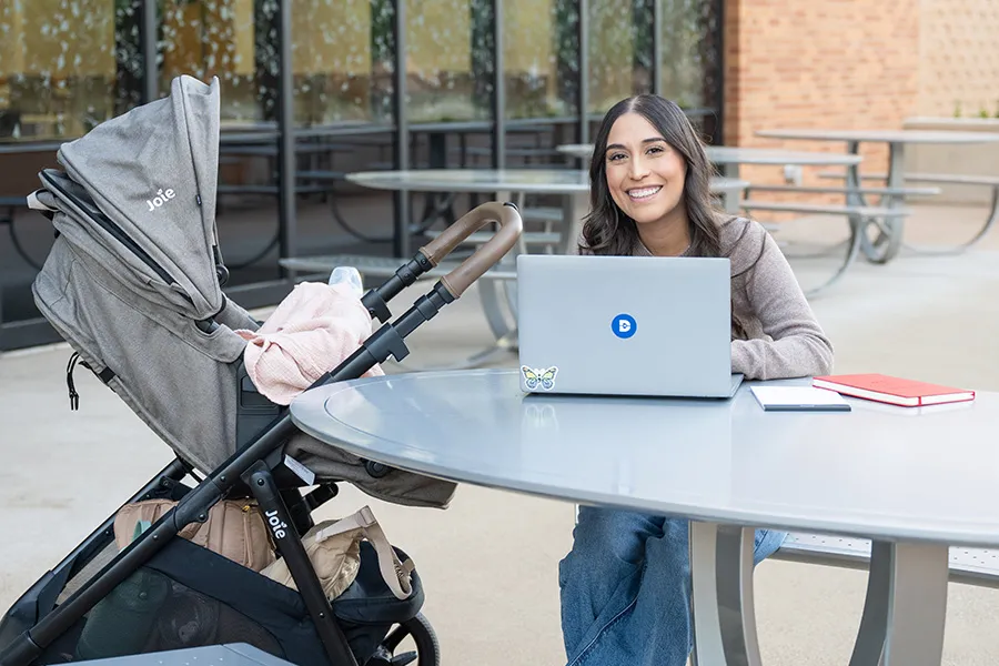 A student sits at a laptop with a baby in a stroller beside her.