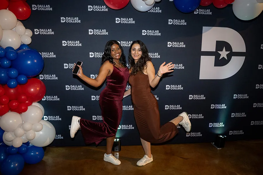 Two students in formal dresses and sneakers stand in front of a Dallas College backdrop at the Sneaker Ball.