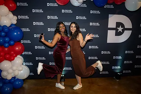 Two students in formal dresses and sneakers stand in front of a Dallas College backdrop at the Sneaker Ball.