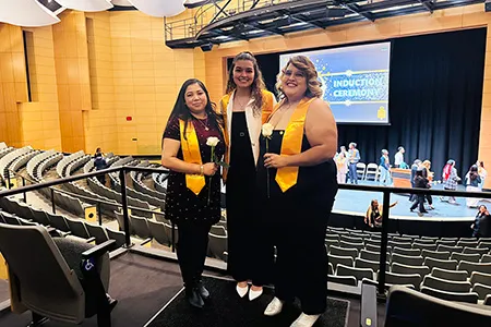 Three students in black dresses standing in front of stage at PTK Induction Ceremony.
