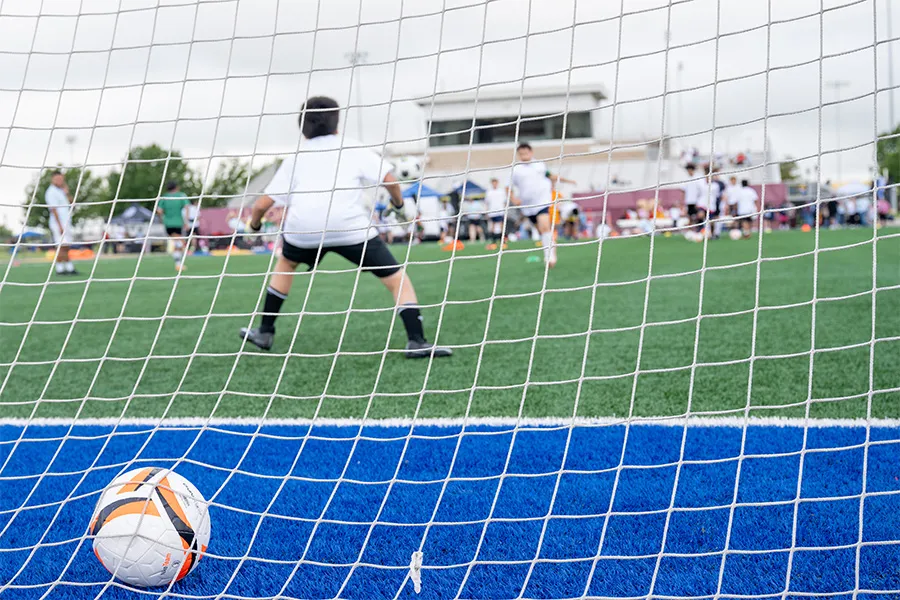 A soccer ball is pictured in a goal with soccer players on the field in the background.