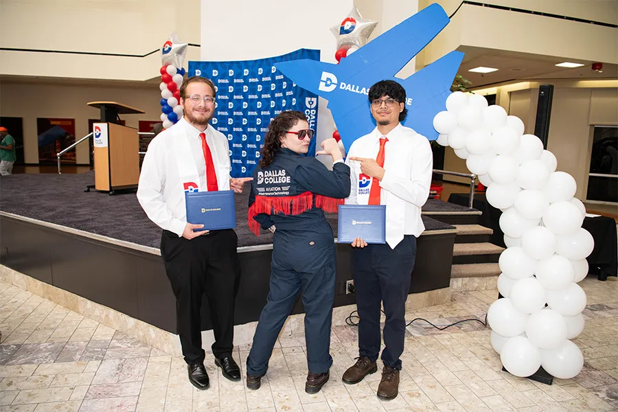 Two AMT graduates stand beside a stage holding their diplomas with a woman wearing a Dallas College jacket in the middle.