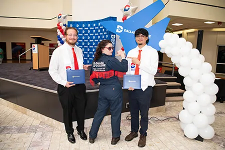 Two AMT graduates stand beside a stage holding their diplomas with a woman wearing a Dallas College jacket in the middle.