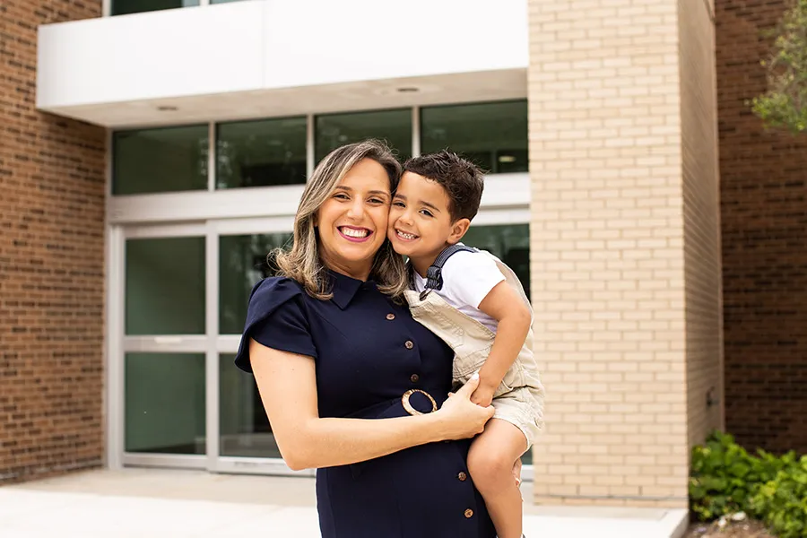 A student-parent stands in front of a building on campus, holding her young child.