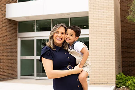 A student-parent stands in front of a building on campus, holding her young child.