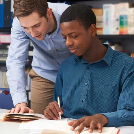 Student writing in a book while receiving guidance from another person in a study environment