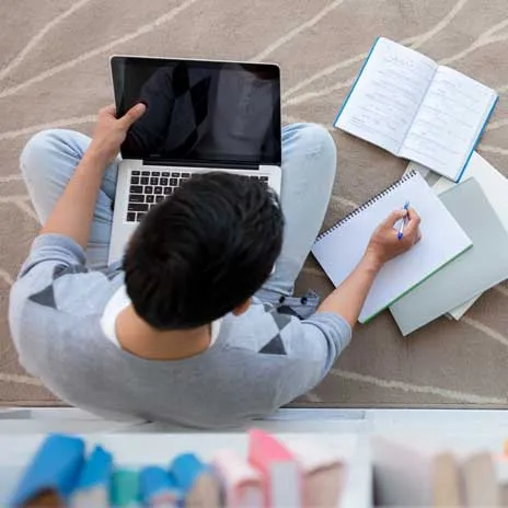 Student sitting on floor using laptop and writing in notebook