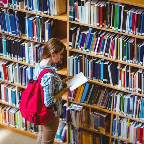 Student with red backpack, reading a book in front of a library bookshelf