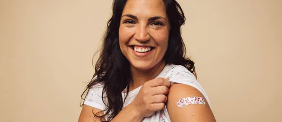 a smiling woman holding up her sleeve showing a bandage on her shoulder