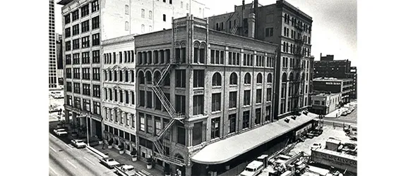 Black and white photo of El Centro Campus building from 1966, featuring a multi-story corner structure with arched windows, an ornate facade, a fire escape, and vintage cars parked along the street.