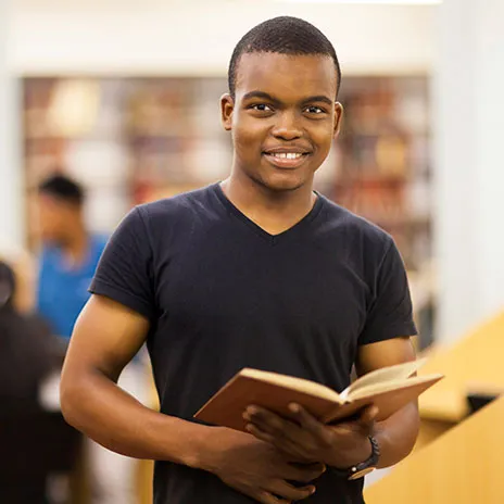 Student holding an open book in a library, with bookshelves and another person in the background