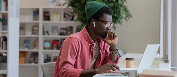 Student wearing a green beanie and red shirt working on a laptop with earphones in and bookshelves in the background.