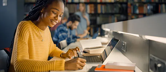 Student in a yellow sweater with braided hair working on a laptop at a library desk.