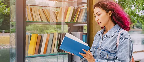 Student with pink hair in a denim jacket reading a blue book in front of a glass bookshelf.