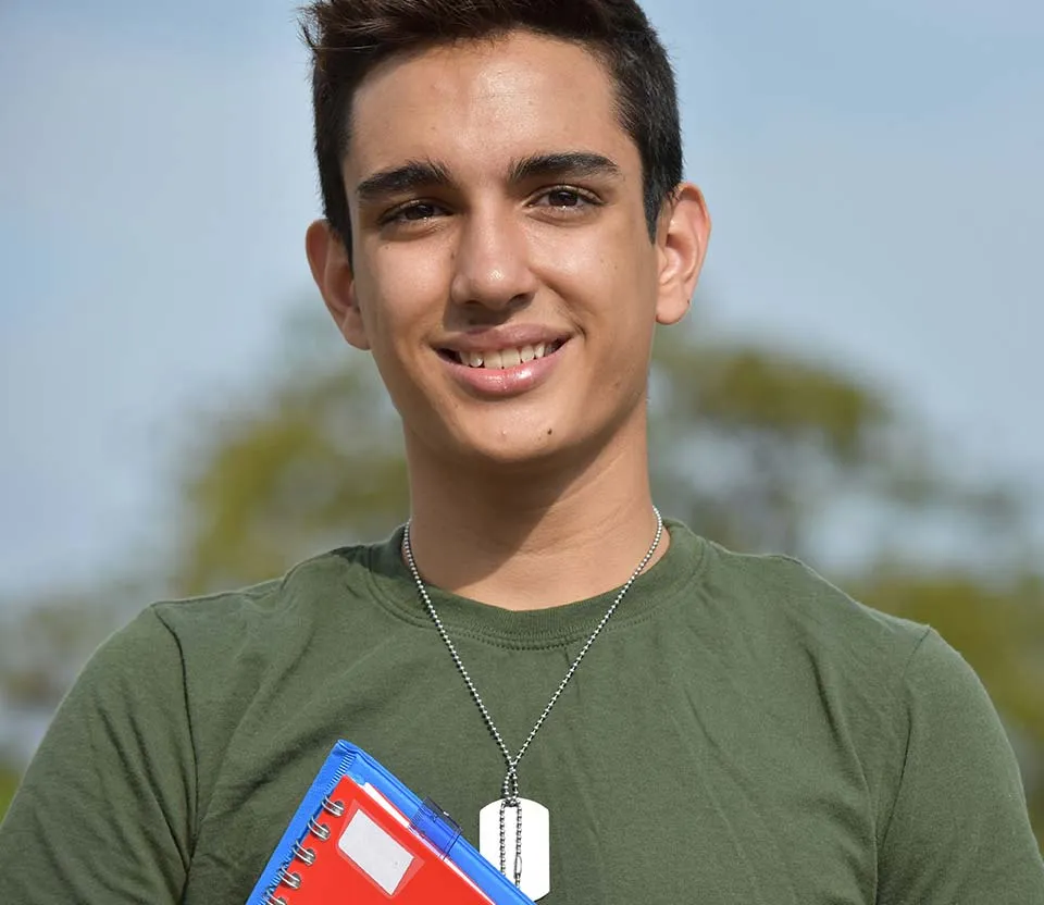 Person in a green shirt with a dog tag necklace holding a red notebook with a blue cover.
