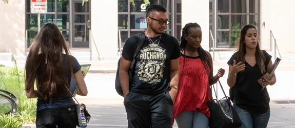 Four students walking on a paved path near a building with large windows; one walks away holding a notebook, while three walk toward the camera talking.