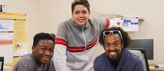 Three students in an office with charts, posters, and computer monitors in the background.