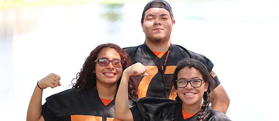 Three students in black and orange sports jerseys posing near a body of water, with two flexing their arms.