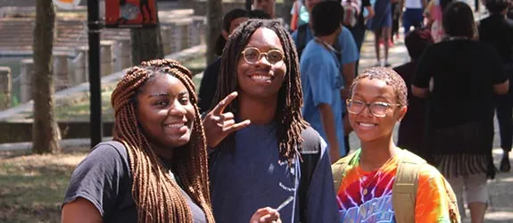 Three students posing for a photo outdoors, surrounded by others walking in a campus with trees in the background.
