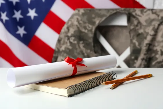 A diploma sits on the table with military jacket draped across a chair and American flag in background.