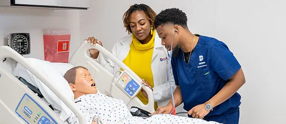 A nursing student checks on a dummy while instructor supervises.