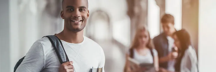 Student with a backpack and books in a hallway, with three others in the background.