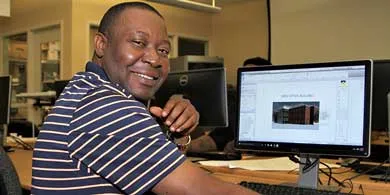 a smiling man in a striped shirt sits at a desk, using a computer
