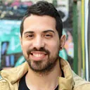 A smiling man with short dark hair and a beard wearing a tan jacket stands outdoors