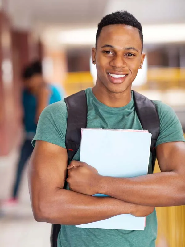 a student smiling in a green shirt with a backpack holds a blue notebook in a bright hallway