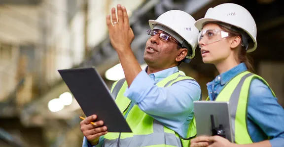 Two construction workers wearing hard hats discuss something while pointing above them