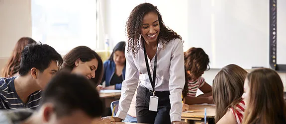 A teacher stands in the middle of a classroom surrounded by the desks of her students