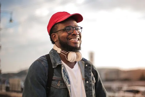 A male student smiles as he walks outdoors, wearing a baseball cap and headphones around his neck