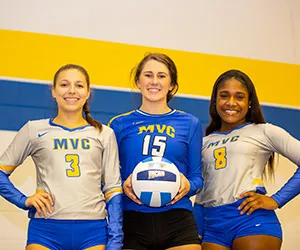 Three Mountain View volleyball players stand together with the center player holding a volleyball