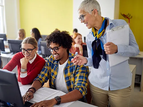 Two students look at a computer in a classroom and the teacher looks over their shoulder