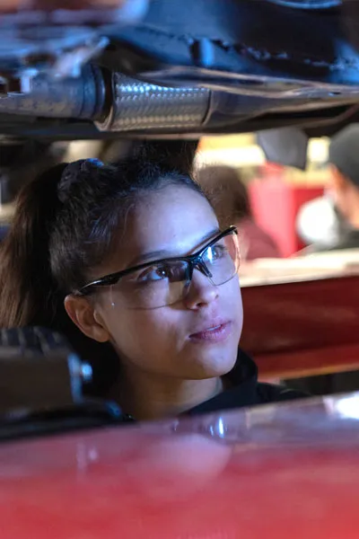 An auto mechanic student looks under a car on a lift.