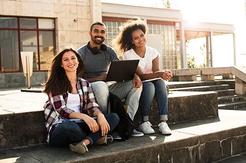 Three students sit outdoors with one student holding a laptop in his lap.
