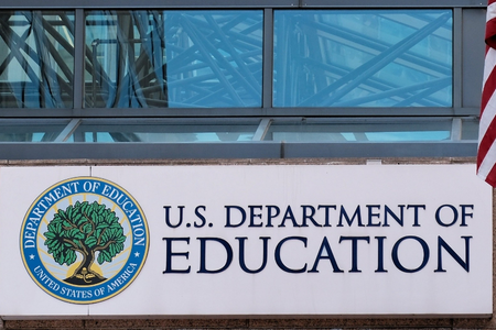 Exterior photo of a U.S. Department of Education building sign with the department seal, next to a U.S. flag.