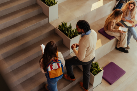 Students sit and talk on tiered indoor steps in a bright, modern academic building, some reading notebooks or tablets, while one student stands on the steps speaking with another; planters and floor cushions are visible nearby.