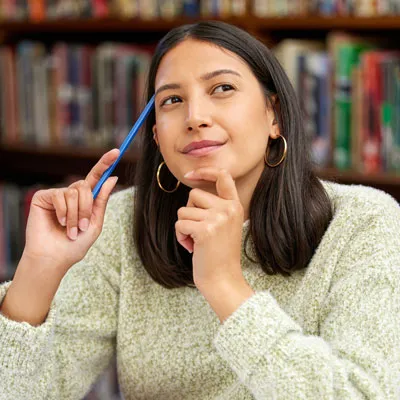 A person sitting in a library holds a pencil upright while resting their other hand near their chin, appearing to think.