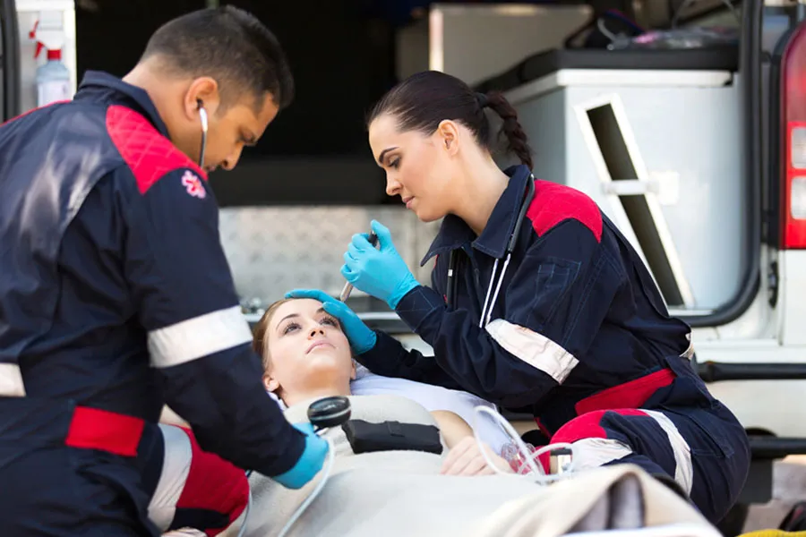 Two emergency medical responders attend to a patient on a stretcher at the back of an open ambulance; faces are blurred for privacy.