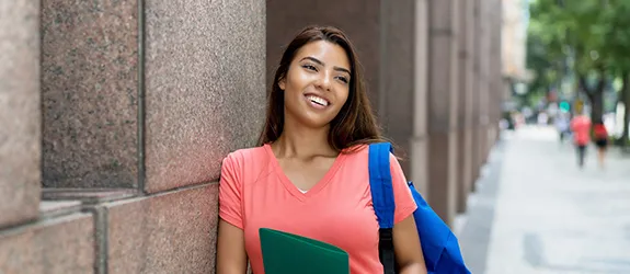 A Latina woman stands with a backpack, leaning against a wall