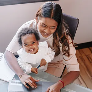 Woman college student working on a laptop with a smiling baby on her lap at a desk