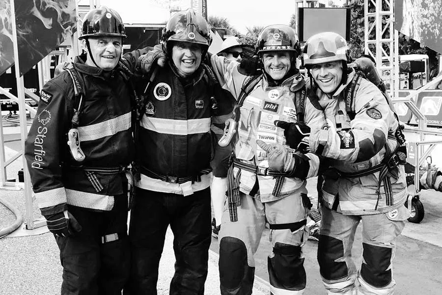 Four firefighters in full protective gear, including helmets and suits, standing close together with arms around each other. The firefighter on the far left has 'Starfield' written on their sleeve. Behind them are firefighting equipment.