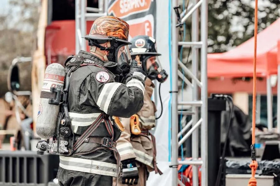 Two firefighters outdoors in full protective gear, including helmets, face masks, and oxygen tanks. The firefighter in the foreground is adjusting their face mask with a gloved hand. Behind them, a metal structure and a red tent are visible.